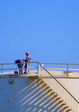 İki mühendis, petrol depolama tankının üstündeki yağ kalitesini dikey çerçevede mavi gökyüzü ile karşılaştırıyor.