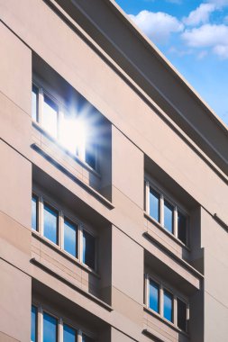 Gri ofis binasındaki birden fazla mavi pencerenin yüzeyinde beyaz buluta ve dikey çerçevedeki mavi gökyüzüne karşı güneş ışığı yansıması