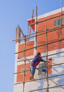 Ahşap iskeledeki Asyalı inşaat işçisi eski evin dışındaki duvarı dikey çerçevede mavi gökyüzüne boyuyor.