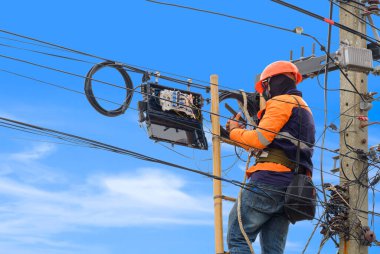 Kablo hatlarının veri numaralarını kontrol etmek için akıllı telefonu kullanan ahşap merdivendeki teknisyen elektrik direğindeki internet dağıtıcı kutusuna fiber optik sistem yerleştirirken mavi gökyüzü arkaplanına karşı 