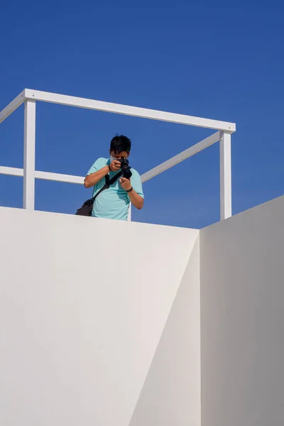 年轻的亚洲男子用数码相机在白色建筑物的屋顶上与蓝天垂直相映成趣 — 图库照片