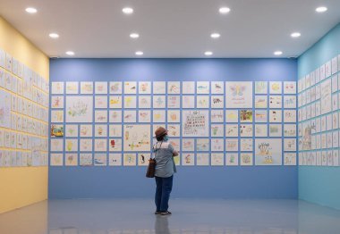 Bangkok, Tayland - 22 Nisan 2022: Asyalı Yetişkin Hippi kadının Bangkok Sanat ve Kültür Merkezi bünyesindeki ilköğretim öğrencileri sergisinin renkli kalem resimlerine bakması