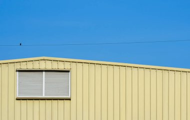 Sarı kıvrımlı çelik depo duvarında alüminyum hoparlör ve mavi gökyüzü arka planında elektrik hattına tünemiş küçük bir kuş.