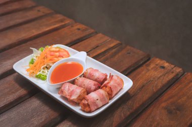 Fried Bacon wrapped sausage rolls with chili sauce and vegetable on wooden table with loft concrete wall in vintage tone style