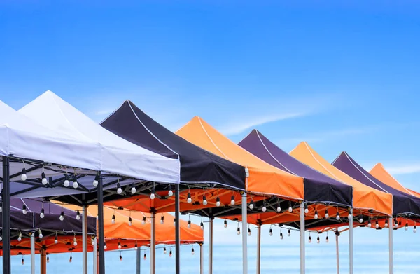 Rows of many foldable canopy tents with hanging LED light bulbs decoration for outdoors event on the beach against sea view background