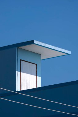 Mavi evin beyaz ahşap çatı kapısının yüzeyindeki güneş ışığı ve gölge dikey çerçevede mavi gökyüzüne karşı, sokak mimarisi arka planda