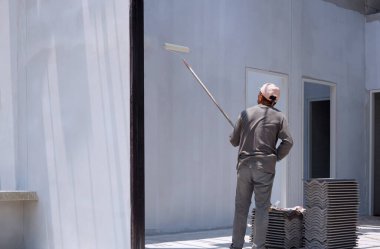 Asyalı inşaat işçisi, inşaat alanının içindeki beton duvara astar beyaz boya sürmek için uzun saplı paten fırçası kullanıyor.