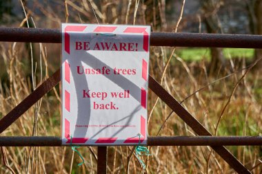 Red and white sign tied to a field gate warning of unsafe trees after the devastation of a big storm