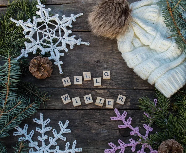 crochet snowflake ornaments on wooden ground with winter slogans and fir branch and cones and a knitted bobble hat background