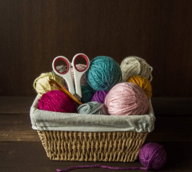 Koyu kahverengi ahşap arka planda, ahşap bir sepetin içindeki ahşap kancalı renkli yün toplar.