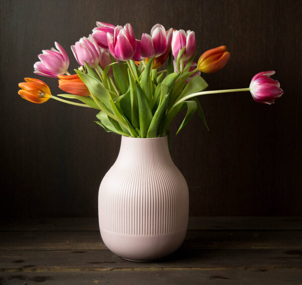 pink and orange colored tulips in vase in front of a dark brown wooden wall background