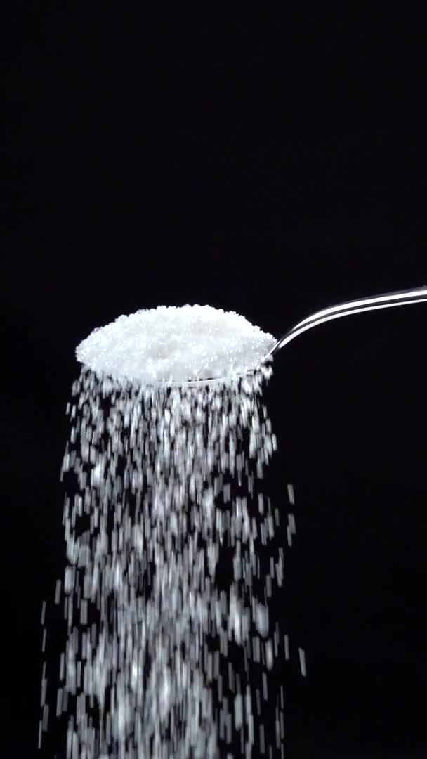 Azúcar cayendo de la cuchara en cámara lenta sobre fondo negro. Vídeo vertical — Vídeos de Stock