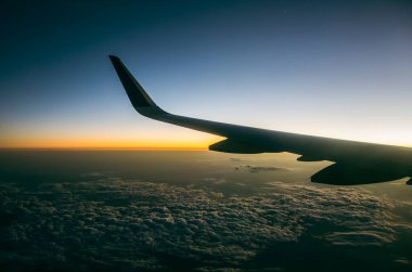 Ufukta gün ışığı kaybolurken uçağın kanadı görünür bulutların üstünde.