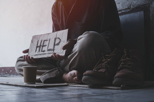 Homeless adult man sitting on the street in the shadow of the building and begging for help and money with sign.  Homeless concept