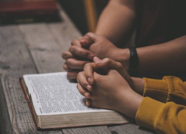 Yakın plan iki Hıristiyan İncil okuyor. Ellerin, kutsal bir İncil 'in üzerinde, ahşap bir masanın üzerinde dua ederken bükülmüş. Çevrimiçi grup ibadeti, Dünya Dua Günü, Uluslararası Dua Günü, Umut, Minnet, Müteşekkir, Güven