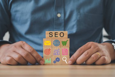 Ellerinde büyüteç simgesi ve alfabe kısaltması SEO olan tahta küpler var. SEO, Arama motoru optimizasyon sıralama konsepti. Trafiği web sitesine tanıtma amaçlı dijital pazarlama stratejisi.
