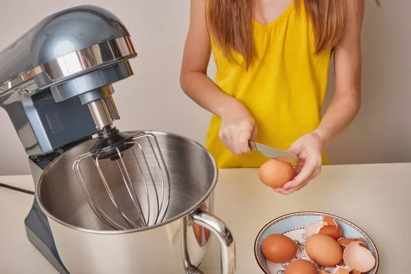 A girl cooks homemade cake in the kitchen, beats eggs in a mixer on the kitchen table. Hands break eggs. Against the background of a white-gray wall. close-up.