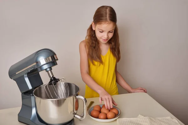 A positive girl of 9-10 years cooks homemade cake in the kitchen, beats eggs in a mixer on the kitchen table. Against the background of a white-gray wall. close-up.