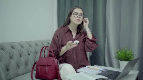 Woman in eyeglasses put on earbuds to make a online call, home interior — Stock Video