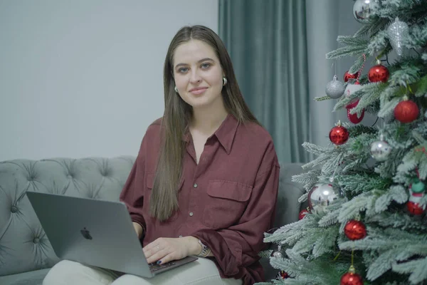 Dizüstü bilgisayarı olan serbest çalışan kadın gülümsüyor. Oturma odasında yeni yıl ağacı