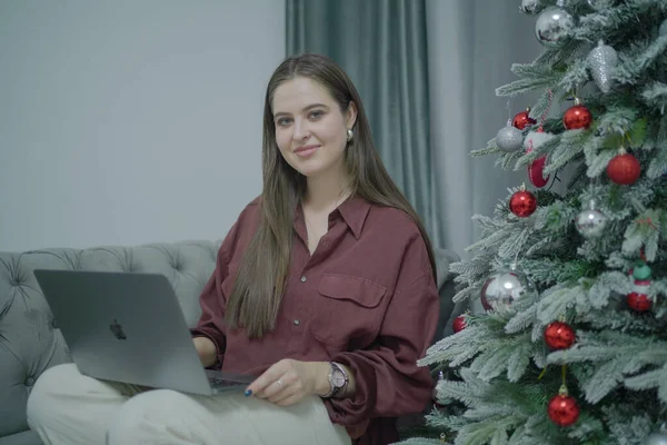 Dizüstü bilgisayarı olan serbest çalışan kadın gülümsüyor. Oturma odasında yeni yıl ağacı