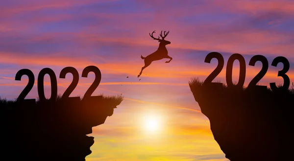 Welcome merry Christmas and Happy new year in 2023. Silhouette Deer jumping across the gap from 2022 to 2023 cliff with Sunset and Twilight Sky background.