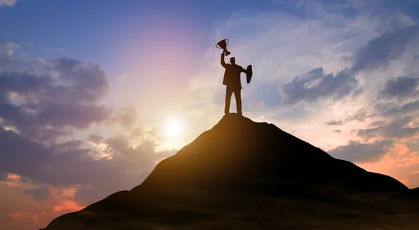 Silhouette victory businessman with trophy climbed to the top of mountain over the sky and sun light background. Success, Achieving goal, Leadership, Career, Winner, and Business achievement Concept.