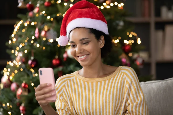 Leende ung afrikansk amerikansk kvinna använder mobiltelefon nära julgran. — Stockfoto