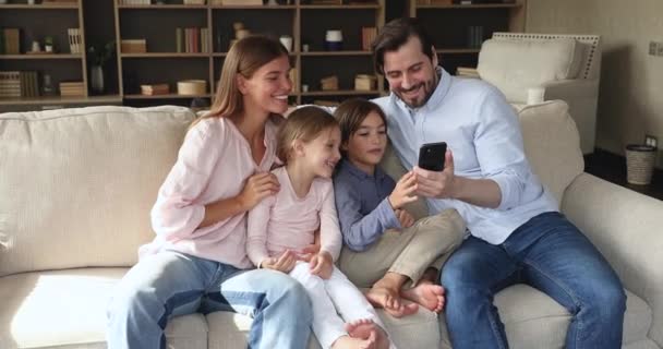 Οικογενειακό ζευγάρι με δύο παιδιά αγκαλιάζονται στον καναπέ χρησιμοποιώντας το τηλέφωνο — Αρχείο Βίντεο