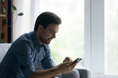 Yandan bakan, gülümseyen, akıllı telefon kullanan, rahat koltukta oturan adam.