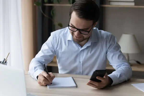 Hombre de negocios sonriente en gafas tomando notas en el cuaderno, sosteniendo el teléfono inteligente — Foto de Stock