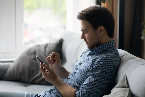 Vista lateral enfocado hombre usando teléfono inteligente, sentado en el sofá — Foto de Stock