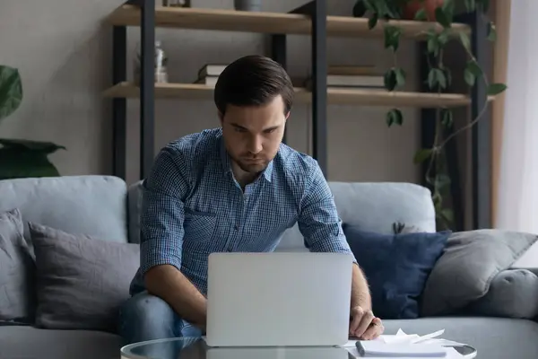 Uomo serio che utilizza il computer portatile, che lavora online, seduto sul divano — Foto Stock