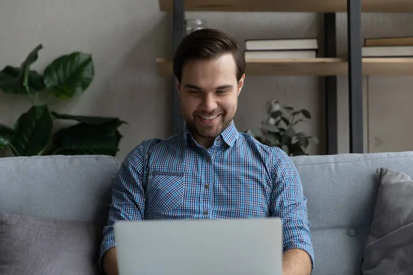 用笔记本电脑微笑的男人，坐在家里的沙发上 — 图库照片