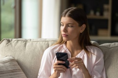 Akıllı telefon tutan, koltukta oturan düşünceli genç bir kadın.