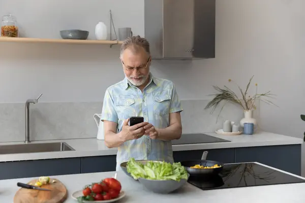 Joyful older mature man using cellphone, preparing food. — Stock Photo, Image