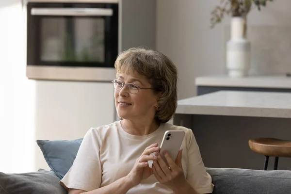 Мечтательная старая женщина средних лет в очках держит мобильный телефон. — стоковое фото