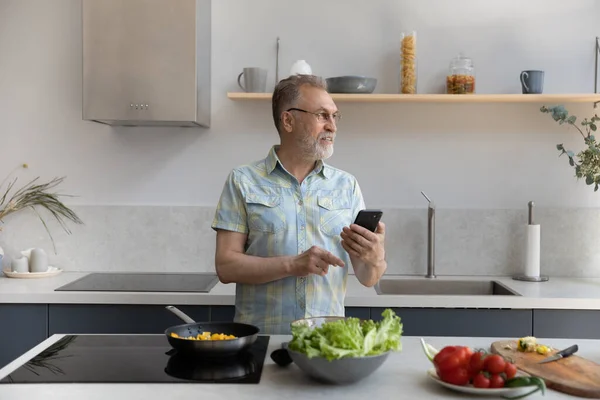 Souriant homme d'âge moyen rêveur utilisant un téléphone portable, cuisine dans la cuisine. — Photo