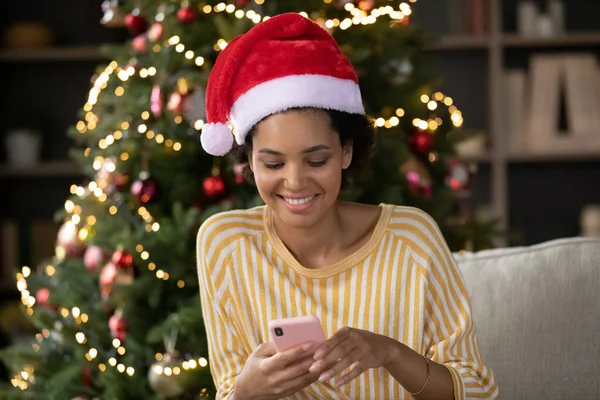 Gelukkig jong Afrikaans amerikaans vrouw met behulp van mobiele telefoon in de buurt van kerstboom. — Stockfoto