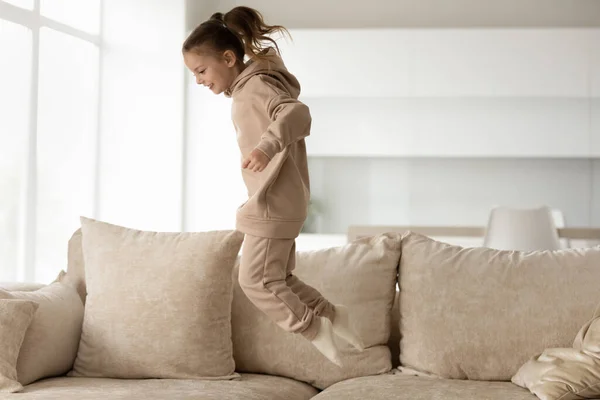 Joyful fetiță copil în loungewear confortabil sărind pe canapea. — Fotografie, imagine de stoc