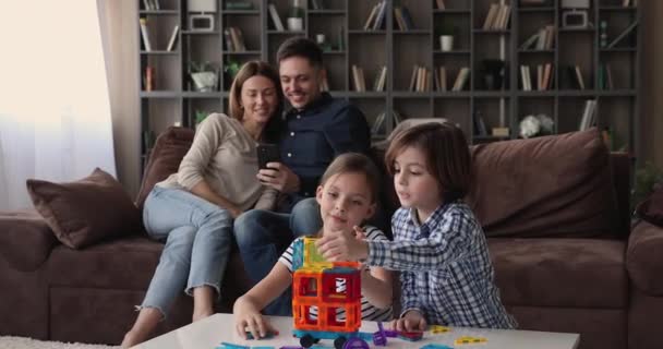 Bambini piccoli che giocano ai giocattoli mentre i genitori usano il cellulare. — Video Stock