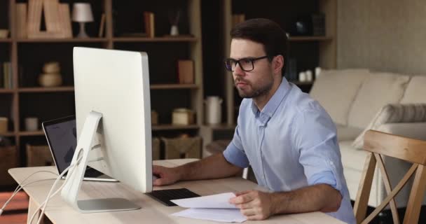 Seriös ung man arbete från hemmakontoret med hjälp av två datorer — Stockvideo