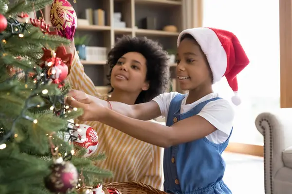 Glückliches kleines afrikanisches Mädchen schmückt Weihnachtsbaum mit Mama. — Stockfoto
