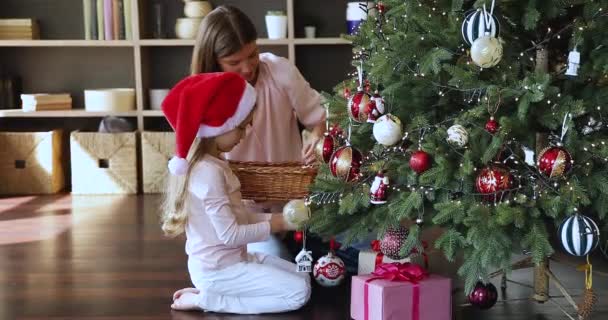 Маленькая дочь сидит на полу рядом с мамой украшать елку — стоковое видео