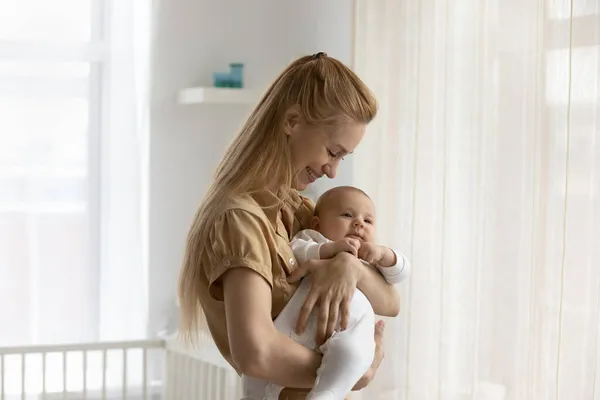 Улыбаясь, любящая молодая мать держит ребенка, стоя дома — стоковое фото
