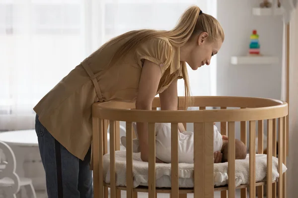 Loving young mother putting sleeping baby in wooden crib — Stock Photo, Image