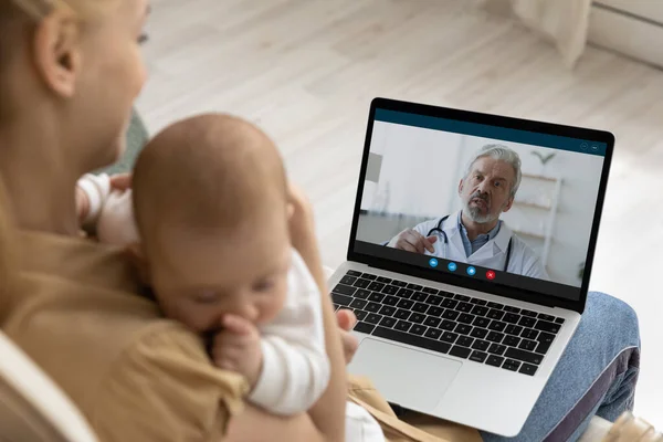 Madre sosteniendo al bebé, usando una computadora portátil, haciendo videollamadas al pediatra —  Fotos de Stock