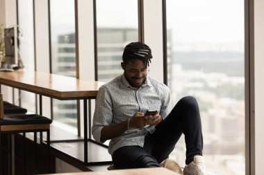 Rahat Afrikalı etnik adam akıllı telefon kullanıyor, pencere kenarında oturuyor..