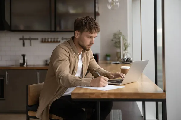 Geconcentreerde jongeman die aan de computer werkt, notities schrijft. — Stockfoto