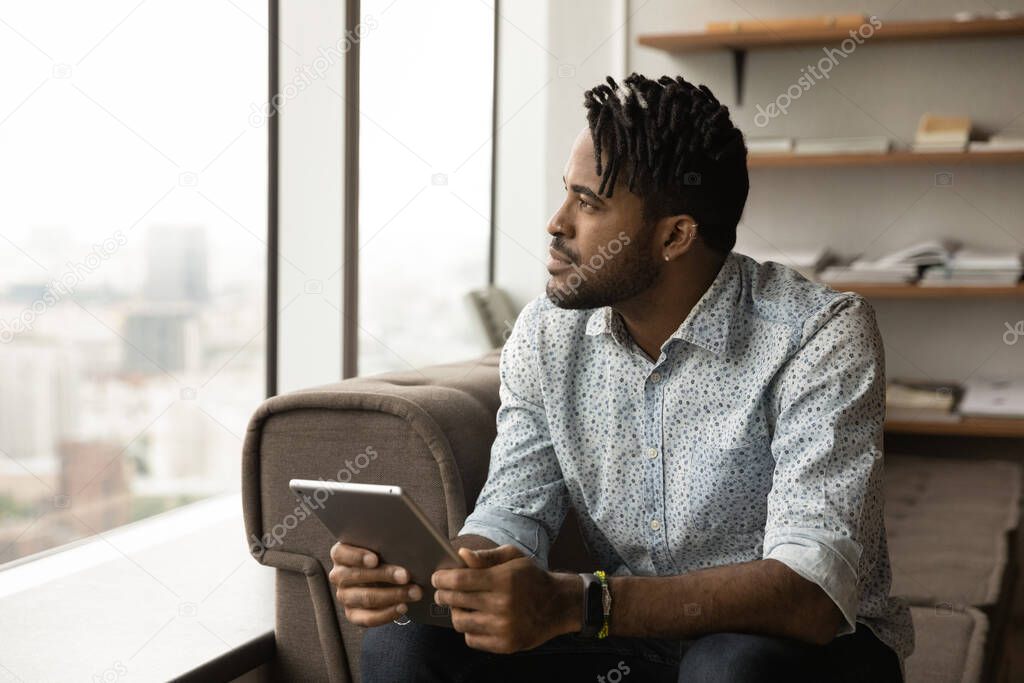 Dreamy thoughtful young african man holding digital tablet.